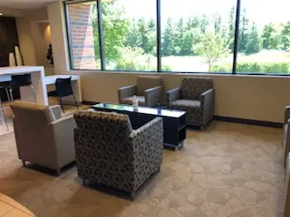 An image of a waiting room, with four brown chairs facing each other with a window in th background.