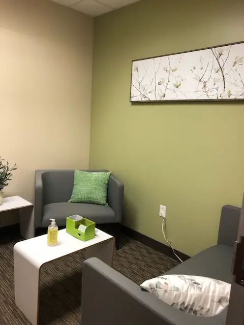 An image of Kate's therapy office, with a gray chair facing a gray couch. The furniture has sitting pillows. There is a small coffee table with tissues and hand sanitizer. A green accent wall features art with a nature scape.